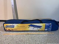 Broadstone folding cot inside blue carrying bag with label showing brand, image of cot, and product details.