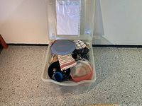 Clear plastic Rubbermaid storage container with lid open showing assorted camping kitchen items including plates, mugs, bowls, pots, pans, lids, utensils, and dish towels.