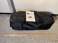 Black carry bag with handles and a ruler to show size, containing the tent and shelter panel with instruction booklet on top.