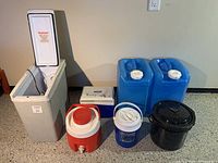 Wide view showing the Koolatron cooler, two large blue water jugs, red and white Thermos jug, blue Hardees jug, black enamel pot, and small Coleman cooler.