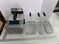 Cuisinart deep fryer next to three frying baskets of different sizes on a white surface, showing overall lot items.