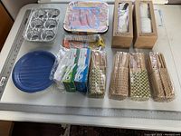 Photo of all disposable kitchenware items including aluminum trays, paper plates, cups, chopsticks, and utensils arranged on a white table.