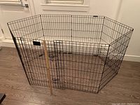 Hexagonal portable metal wire kennel with door and locking latches, placed on wood floor.