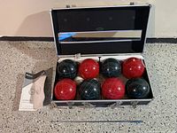 Open case showing 4 red bocce balls, 4 grey bocce balls with grid lines, and one white ball. Instruction booklet and metal pointer shown beside case.