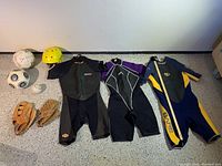 Overview of all lot items including three wetsuits, two soccer balls, softball, two baseball gloves, and yellow helmet