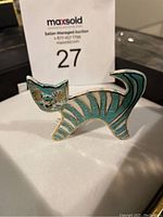 Front view of enamelled ceramic cat brooch showing detailed face, stripes and tail, placed on a white surface with auction tag behind.