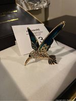 Front and side view of a vintage bird brooch with teal blue enamel and clear rhinestones on a gold-tone base, placed on a white surface with MaxSold lot number paper in the background.
