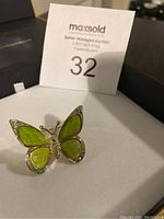 Green and gold butterfly brooch with light green wings accented with small green and clear rhinestones lying on a white surface with auction lot number in background.