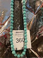 Photo of a round turquoise stone necklace displayed on a black jewelry stand with reflective silver background and a lot tag.