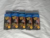 Five unopened packs of Black Flag Fly Paper strips lined up on a white cloth background.