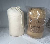 Two sets of sheets, one beige in a drawstring cloth bag, one gold wrapped in plastic with Merit Linens branding.