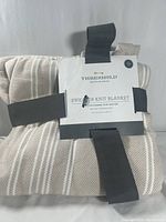 Folded Threshold Sweater Knit Blanket with label and black elastic bands, showing beige color with white stripes.