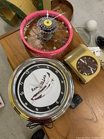 Three clocks on table: vintage Bankvalab electric wall clock, pink bicycle wheel clock with colorful beads, yellow plastic mantle clock