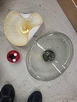 Three assorted glass items on a floor: large clear-grey bowl with central depression, white-yellow ruffled bowl, and small red spherical glass item with black stars inside