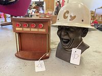 Photo showing teak cocktail dispenser with four red knobs and attached tubes alongside sculpture bust wearing "Cy" helmet