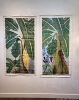 Two full oil paintings featuring banana trees hung side by side on a white wall with a wooden floor and table in frame.