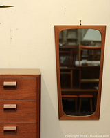 Frontal view of trapezoid teak framed mirror hanging on wall beside furniture