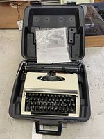 Vintage cream-colored manual typewriter with black keys inside a black hard plastic carrying case with the instruction manual on top.