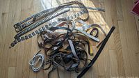 Wide top-down view of a pile of various belts on a wooden floor showing metal chain belts, leather belts, and braided belts.