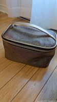 Rectangular metallic cosmetic bag on wooden floor, closed, showing textured surface and top handle.