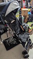 Side view of small black and grey single seat stroller showing fabric seat, canopy, wheels, and metal frame.