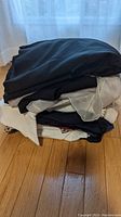 Stack of folded women's clothing in black, white, beige and dark colors on wooden floor near curtain.