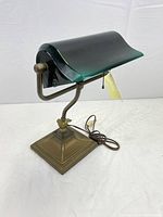 Bankers desk lamp showing heavy brass base, adjustable arm and green glass shade with pull chain switch and brown cord.