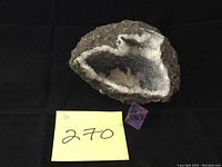 Geode with visible white and gray crystal interior alongside a small purple crystal piece on a black background with a yellow note showing lot number 270.