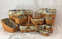 Photo showing all 10 hand woven Longaberger baskets arranged with visible Rose Trellis patterned liners inside each basket. Baskets vary in size and shape including oval, round, and rectangular.