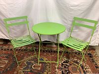 Front view of two bright green folding metal chairs and round table set on a patterned rug, washable surface but showing some wear.