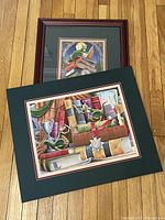 Two Randal Spangler dragon prints: one framed print showing a dragon on a stack of books with trophies and medals, and one matted print with a dragon on books under a moonlit window.