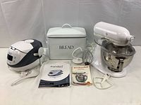 Wide shot of all items: white KitchenAid stand mixer, Panda Ninja rice cooker with paddle, painted metal bread box and instruction manuals