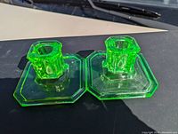 Pair of fluorescent green Vaseline glass Art Deco candlesticks with octagonal bases placed side by side on a flat surface, showing front and side views under natural sunlight.