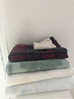 Stack of five towels. Bottom three are bath towels in white and light blue shades. Fourth towel is maroon and grey checkered bath towel. Top towel is small white face towel.