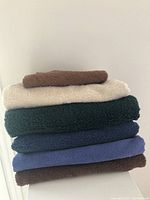 Stacked towels showing five bath towels in cream, dark green, navy blue, blue, and brown colors, plus one smaller brown hand towel on top.
