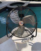 Black Climate Keeper high velocity fan positioned outdoors on a white stool near a pool. Image shows the front protective grill, metal blades, and frame with power cord wrapped.