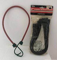 Photo showing one red and black 18-inch bungee cord with green hooks along with pack of four black rubber tie-down straps with metal S-hooks in original packaging.