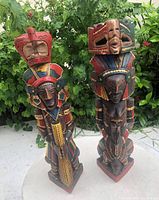 Front view of two hand-carved wooden statues with detailed human figurative designs and colorful paintwork in natural outdoor background.