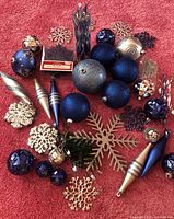 Top view of various blue and silver Christmas ornaments arranged on a red carpet surface. Includes baubles in shiny, matte, and faceted finishes, multiple snowflake shapes in metallic colors, elongated teardrop shapes, and a decorative candle light.