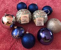 Blue and silver Christmas tree ornaments arranged on red carpet, showing various sizes, finishes, and decorative details including glitter, matte blue, green gems, and gold leaf patterns.