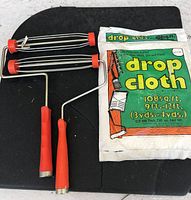 Photo of two metal paint roller frames with red handles and a packaged drop cloth for painting, showing size and packaging details on the drop cloth.