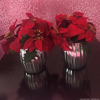 Pair of two artificial red poinsettia flower arrangements in silver metallic ribbed vases placed on a black surface in front of a sparkly pink background.