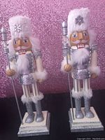 Photo of two Christmas nutcracker soldier figurines standing side by side, both with silver and white glitter attire, fluffy white hats and beards, and holding silver staffs.