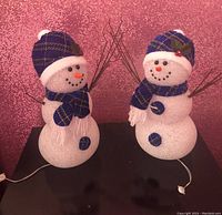 Pair of battery-operated Christmas snowmen on dark surface with pink glitter background. Each snowman dressed in blue and green plaid hat and scarf, stick arms, and carrot nose. White textured finish and round body sections visible.