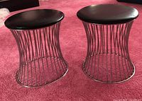 Pair of faux black leather-top side tables or stools with chrome wire bases shown on pink carpet.