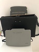 Light gray Polaroid Automatic 103 Land Camera with black carrying case and instruction manual behind it. Manual shows wear at edges.