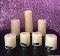 Photo showing set of 7 vanilla scented candles in 3 sizes: 4 short, 2 medium, 1 tall, all wrapped in plastic.