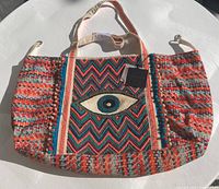 Full view of colorful woven and beaded tote bag with eye motif on center front panel, showing handles, side gathered designs, and original tag.