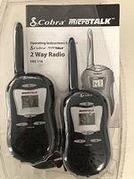 Clear photo showing two black Cobra MicroTALK FRS 110 walkie-talkies with antennas and LCD screens, alongside a printed operating instruction sheet.