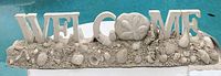 Photo of a white and beige wooden welcome sign with the word 'WELCOME' and decorated with seashells including starfish and sand dollar.